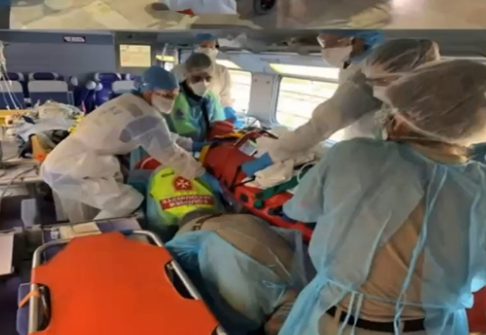 Interior d'un TGV sanitari COVID. Personal sanitari equipat amb bates, mascaretes, casquets i ulleres desplacen un pacient empaquetat en un sac de buit cap a sobre les esquenes d'un parell de socorristes que esperen ajupits per fer de suport