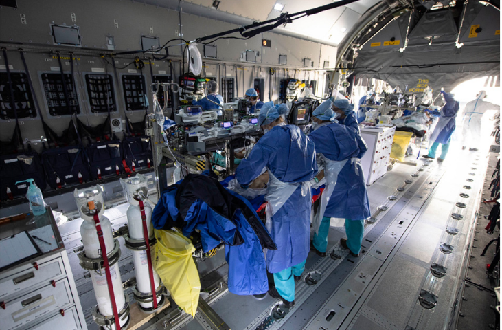 Interior d'un avió de transport militar modificat per a poder traslladar pacients amb COVID. Es veuen diverses lliteres al centre de l'avió amb pacients i equipament mèdic. Una colla de sanitaris amb els EPI COVID envolten les lliteres. Al fons es pot veure la porta posterior de l'avió oberta.